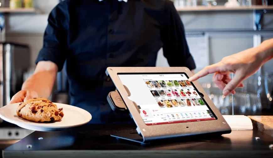 Tablet-based point of sale system in a cafe, with a person serving pastries and another person interacting with the touchscreen.