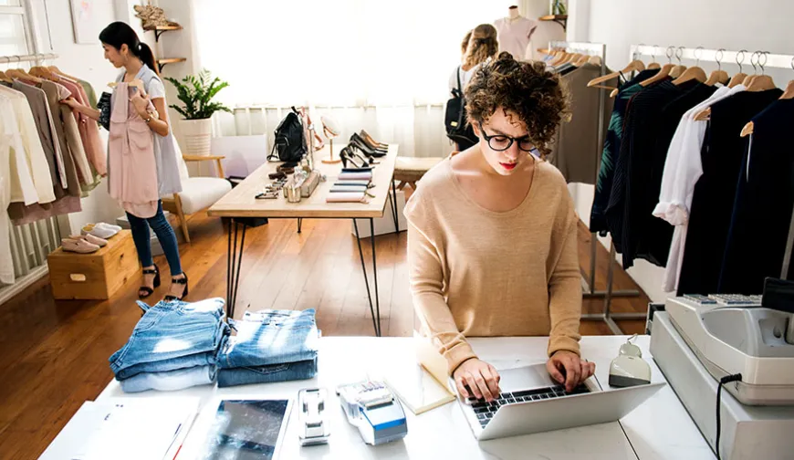 Store employee using a laptop at the point of sale in a clothing boutique, while customers shop in the background.