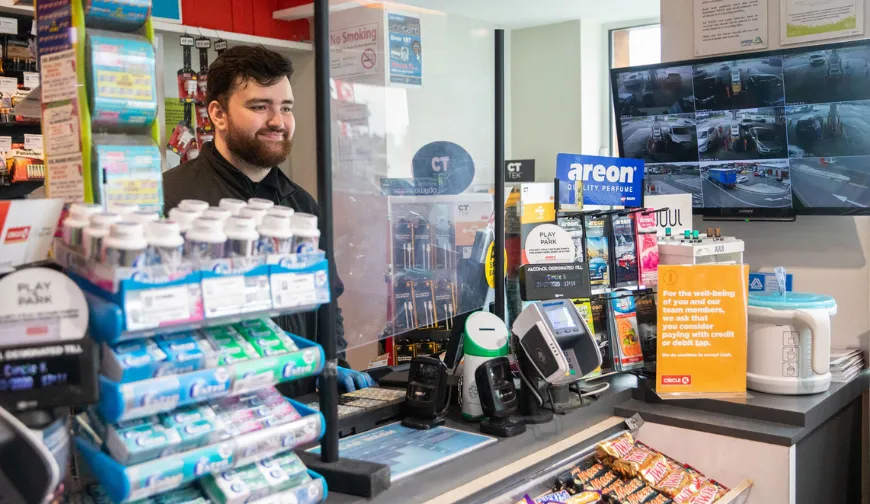 A cashier works at a retail point of sale, surrounded by various products, payment terminals, and surveillance screens.