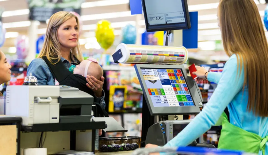 Customer with a baby at a point of sale, interacting with a cashier using a touchscreen checkout system in a supermarket.