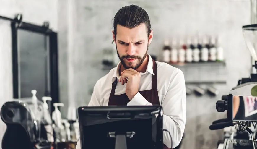 A barista in an apron standing at a point of sale terminal, concentrating on the screen in a coffee shop.